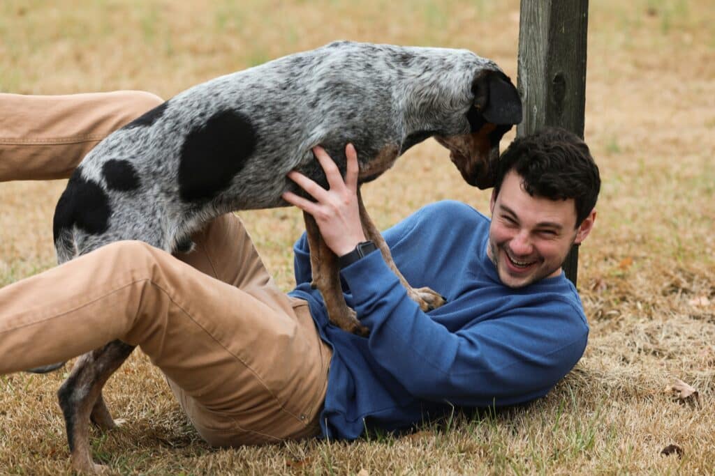 Homem deitado na grama, sorrindo e brincando com seu cachorro preto e branco — atividades para divertir seu mascote sem gastar muito.