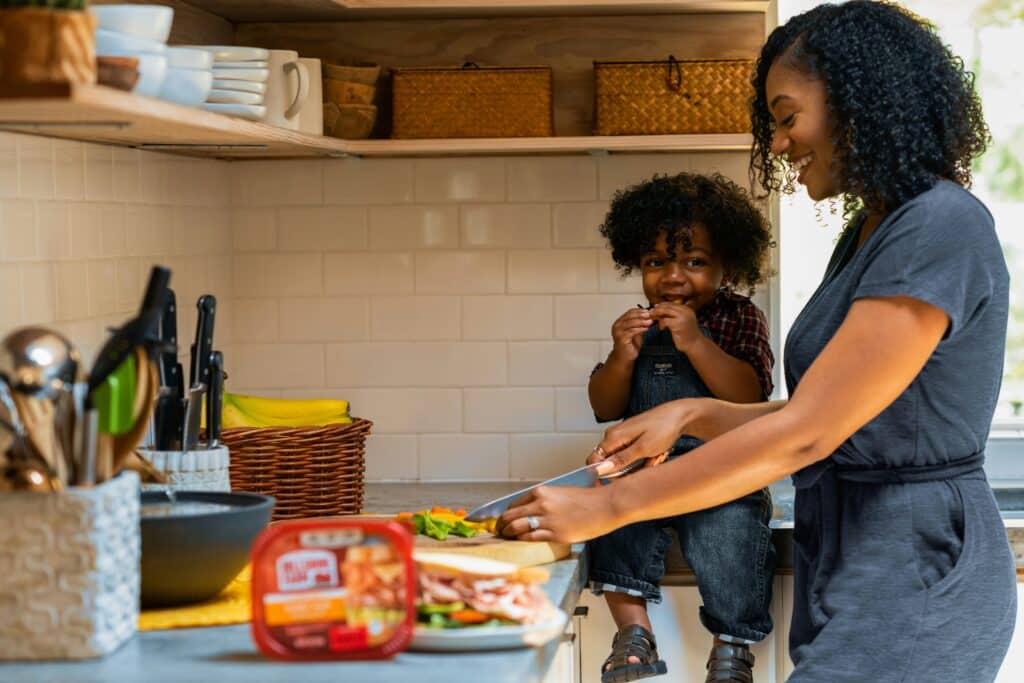 Ideas para hacer la hora de la comida más entretenida: uma mulher corta verduras enquanto seu filho merienda na cozinha.